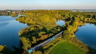 Blau-türkiser Regionalzug fährt entlang eines Sees durch eine grüne Landschaft mit Wäldern und Wiesen unter klarem Himmel, aufgenommen aus der Luft.